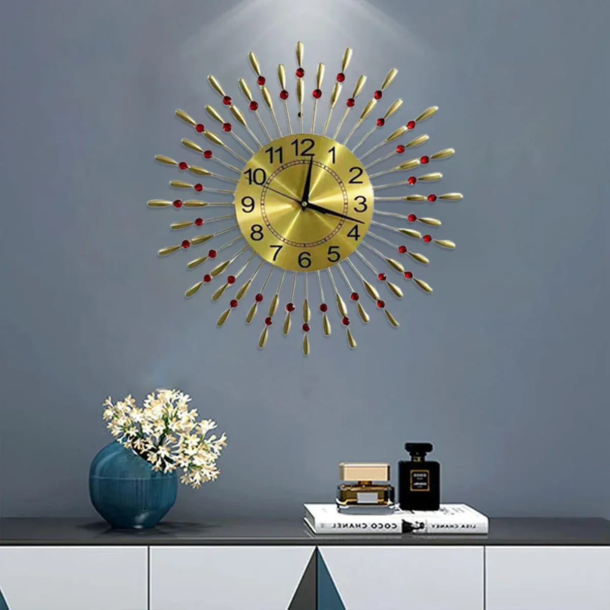 A gold sunburst wall clock with red bead accents is mounted on a gray wall above a console table styled with a blue vase of flowers and a perfume bottle.
