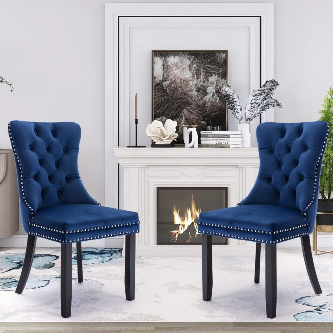 Pair of blue velvet tufted wingback dining chairs with stud trim and solid dark wooden legs, set in a modern living room.