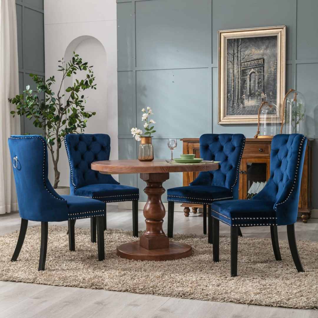Set of four blue velvet tufted wingback dining chairs with stud trim and solid dark wood legs around a round wooden pedestal .