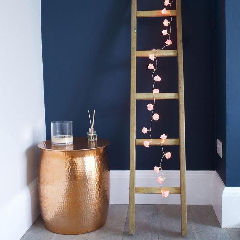 Wooden ladder leaning against navy blue wall with 50 pink velvet rose fairy lights draped vertically, vintage-style gold.