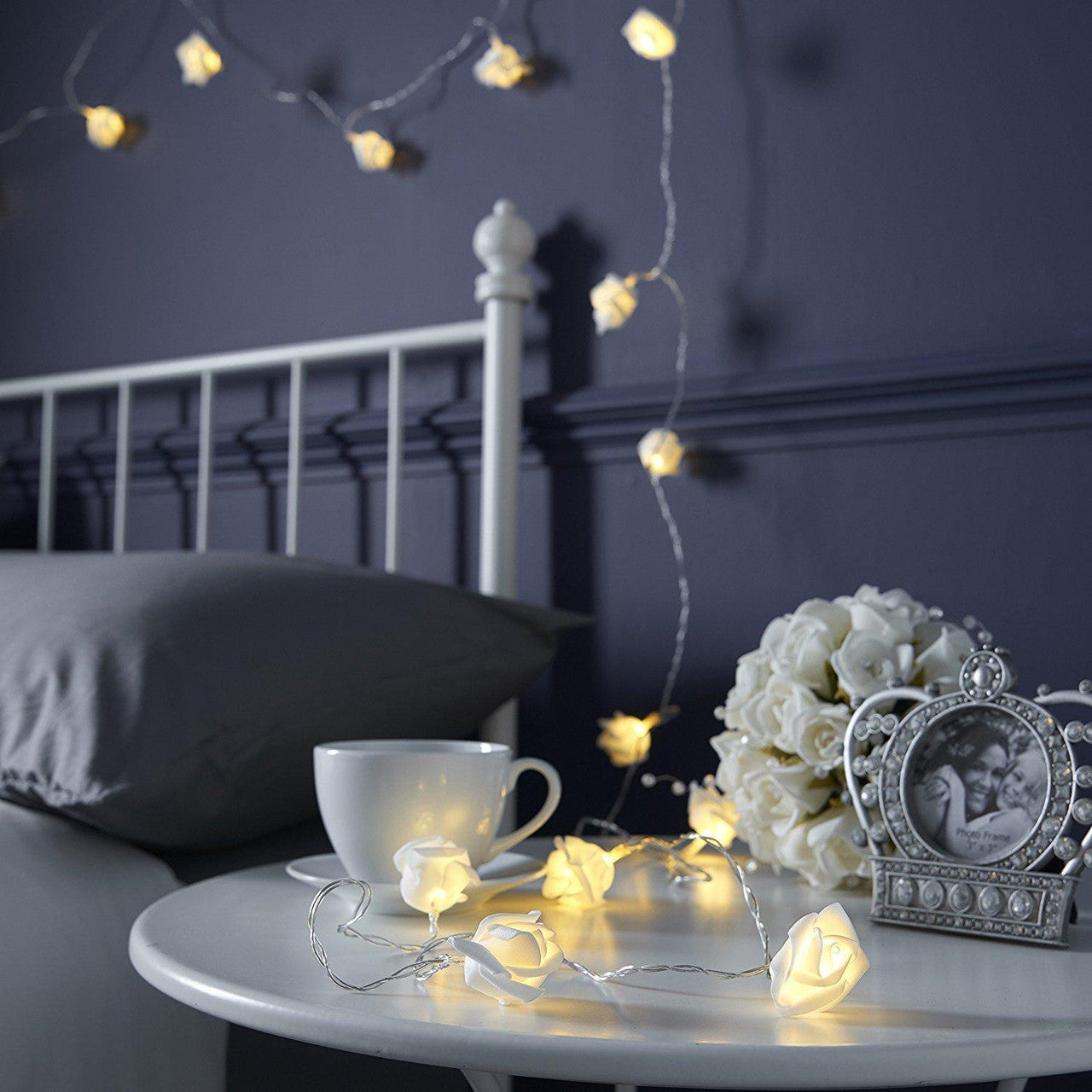 50 white velvet rose 3D fairy lights draped over vintage white metal bedframe and cozy gray bedding in retro-inspired.