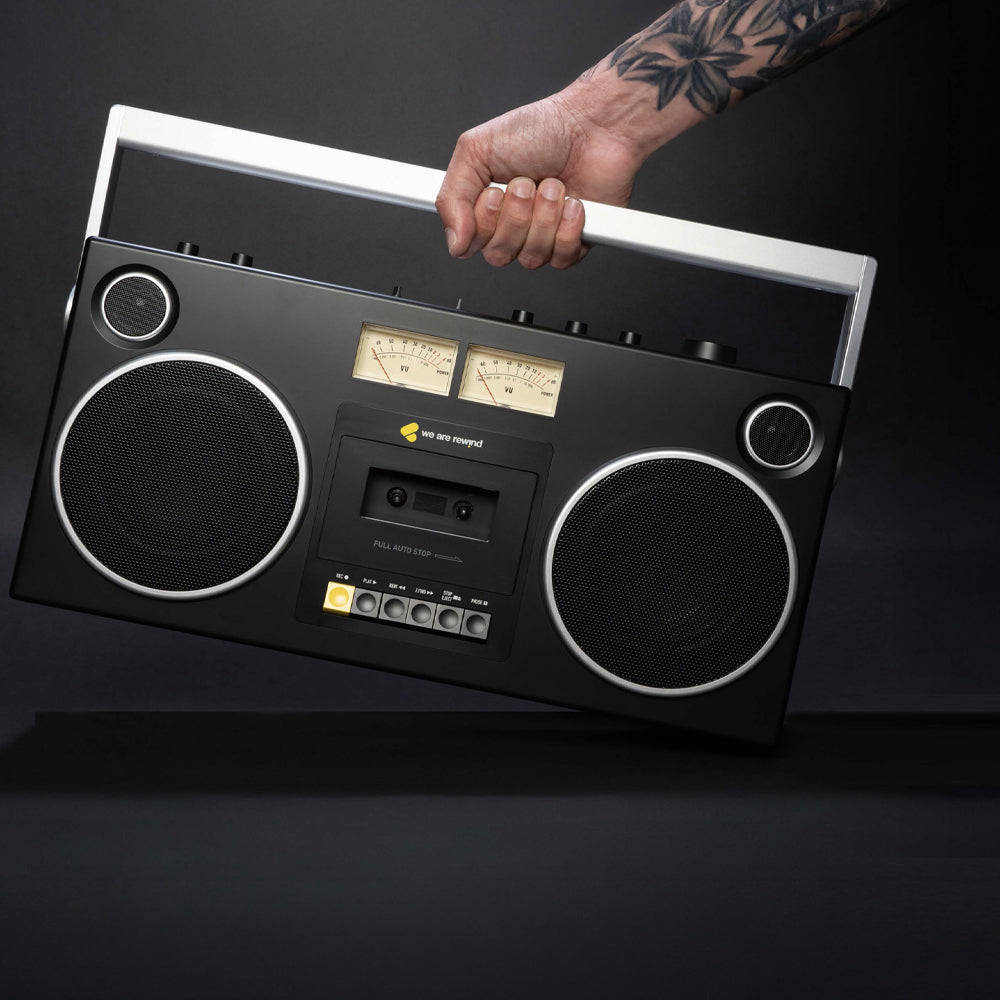Black retro velvet boombox-style cassette player with large speakers and analog meters, held by tattooed hand against dark.