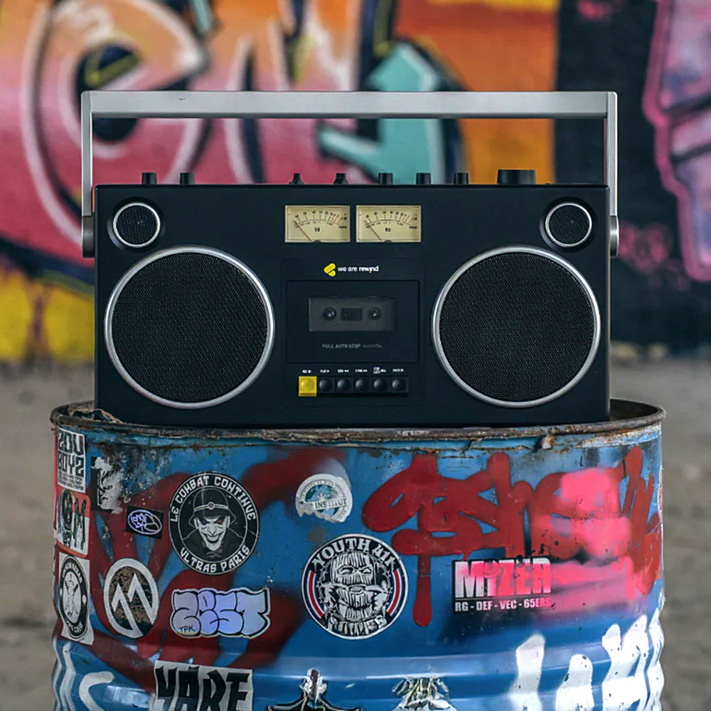 Black retro Getto Blaster cassette player with vintage dials and buttons, sitting on a graffiti-covered barrel against a.