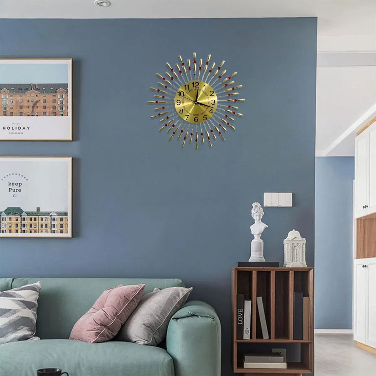 A gold sunburst wall clock with colorful geometric hour markers is mounted on a blue-gray living room wall above a green sofa and next to a wooden side table.