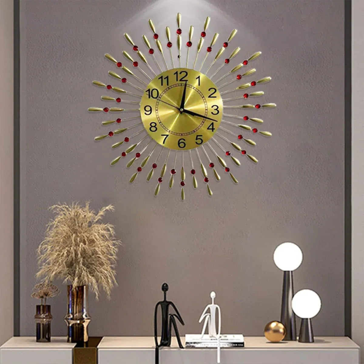 A modern sunburst wall clock with a gold-toned face and radiating metallic spikes accented by small red beads, mounted on a gray wall above a styled console table with decorative objects.