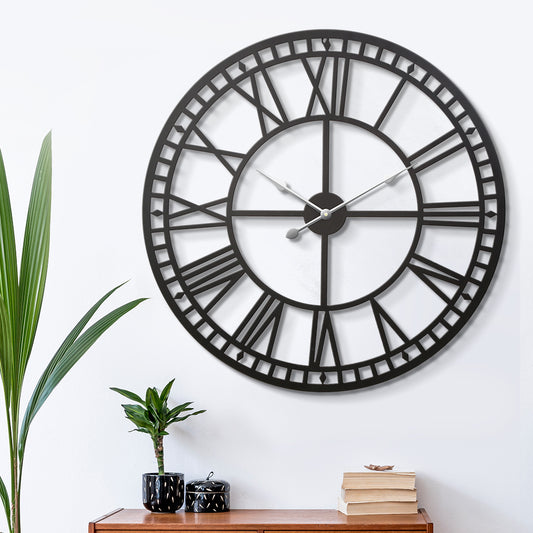 Large black metal wall clock with vintage Roman numerals in a retro style, hanging on a white wall above a wooden surface.